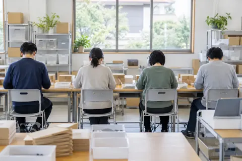 作業台に並んで作業をする人たちの後ろ姿（障害者就労支援・作業所の写真 No.22）