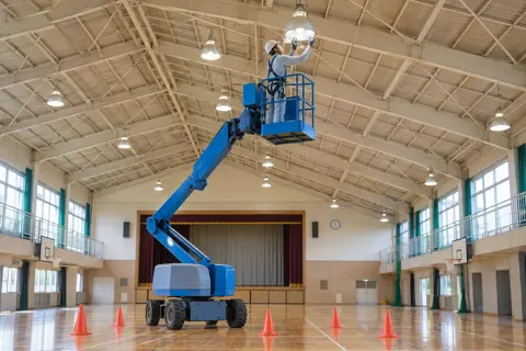 体育館の照明を高所作業車で取りかえる作業員（電気工事・空調・電気設備の写真 No.53）
